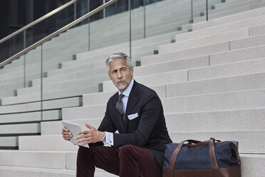 Porträt eines eleganten Geschäftsmannes mit Reisetasche und Tablet auf einer Treppe sitzend - RORF01495