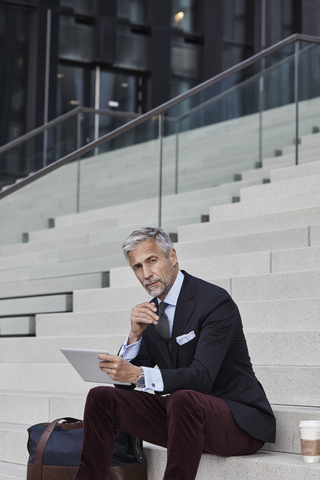 Porträt eines eleganten Geschäftsmannes mit Reisetasche, Coffee to go und Tablet auf einer Treppe sitzend, lizenzfreies Stockfoto