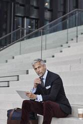 Porträt eines eleganten Geschäftsmannes mit Reisetasche, Coffee to go und Tablet auf einer Treppe sitzend - RORF01496
