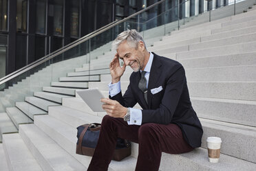 Fashionable businessman with travelling bag and coffee to go sitting on stairs using tablet - RORF01498