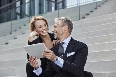 Portrait of two laughing business people with tablet sitting together on stairs - RORF01501