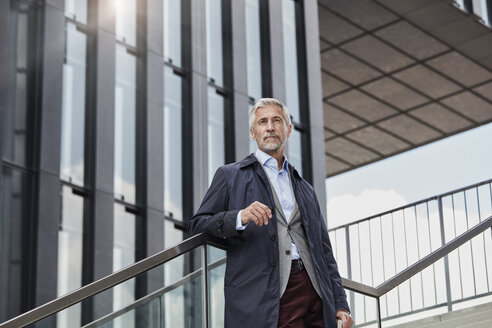 Portrait of mature businessman standing on stairs in front of modern office building looking at distance - RORF01507