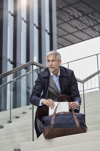 Porträt eines reifen Geschäftsmannes mit Reisetasche und Tablet, der auf einer Treppe im Freien hockt, lizenzfreies Stockfoto