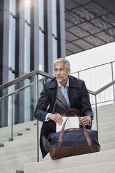 Porträt eines reifen Geschäftsmannes mit Reisetasche und Tablet, der auf einer Treppe im Freien hockt - RORF01508