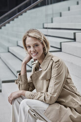 Portrait of smiling blond businesswoman wearing beige trenchcoat sitting on stairs - RORF01522