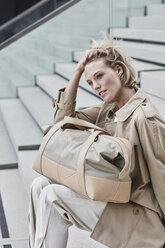 Portrait of blond businesswoman with travelling bag wearing beige trenchcoat sitting on stairs - RORF01524