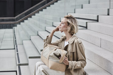 Modische blonde Geschäftsfrau im beigen Trenchcoat auf der Treppe sitzend mit Reisetasche - RORF01525