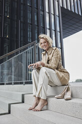 Portrait of laughing young blond woman with digital camera sitting barefoot on stairs - RORF01544