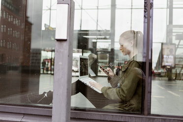 Woman using laptop and smart phone while sitting by window at station - MASF08612