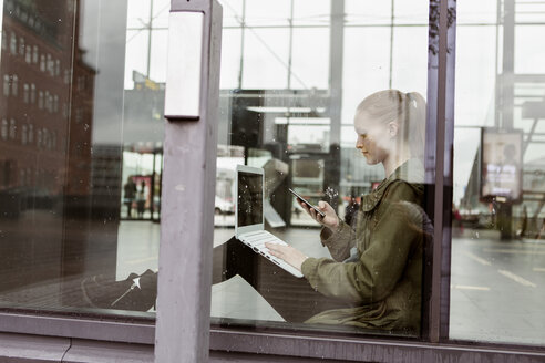 Frau benutzt Laptop und Smartphone, während sie am Fenster des Bahnhofs sitzt - MASF08612