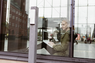 Thoughtful woman using laptop while looking through window at station - MASF08614