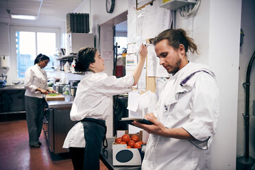 Female chef making checklist while colleagues working in kitchen - MASF08650