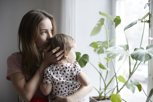 Mother kissing daughter while looking through window at home - MASF08741