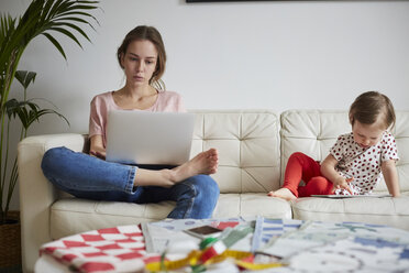 Modedesignerin schaut auf ihren Laptop, während ihre Tochter auf dem Sofa zu Hause ein digitales Tablet benutzt - MASF08749