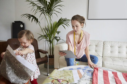 Modedesignerin mit digitalem Tablet, die Textilien untersucht, während ihre Tochter zu Hause spielt - MASF08761