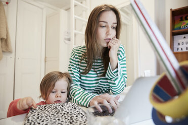 Neugieriges Mädchen, das ein Buch berührt, während die arbeitende Mutter einen Laptop am Tisch benutzt - MASF08772