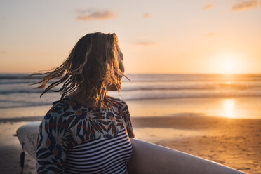 Weiblicher Surfer mit Surfbrett schaut bei Sonnenuntergang am Strand aufs Meer - MASF08784