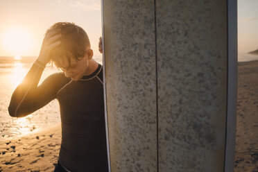 Junger Mann mit Kopf in der Hand, der am Strand bei Sonnenuntergang auf einem Surfbrett steht - MASF08793