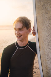 Lächelnder junger Mann mit Surfbrett am Strand bei Sonnenuntergang - MASF08795