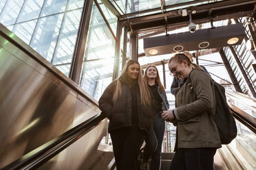 Lächelndes Teenager-Mädchen, das ein Mobiltelefon benutzt, während es mit Freunden auf einer Rolltreppe steht - MASF08806