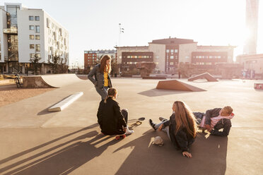 Stehendes Mädchen im Gespräch mit Teenager-Freunden, die in einem Skateboard-Park an einem sonnigen Tag sitzen - MASF08813