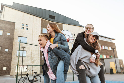 Lächelnder Teenager und Mädchen, die Freunde huckepack nehmen, während sie an einem Gebäude in der Stadt stehen - MASF08823
