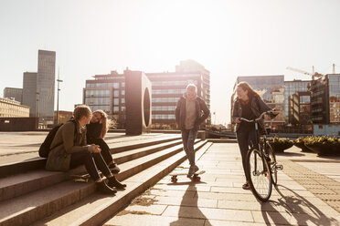 Teenage girl riding bicycle and boy skateboarding while friends sitting on steps in city - MASF08826