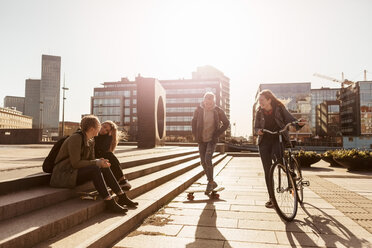 Teenager-Mädchen auf dem Fahrrad und Junge auf dem Skateboard, während Freunde auf einer Treppe in der Stadt sitzen - MASF08826