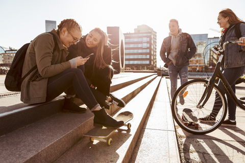 Teenager-Mädchen unterhalten sich auf einer Treppe, während Freunde mit Fahrrad und Skateboard in der Stadt stehen, lizenzfreies Stockfoto
