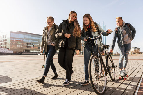 Teenager im Gespräch mit einem Freund, der ein Fahrrad hält, während ein Junge in der Stadt Skateboard fährt - MASF08829