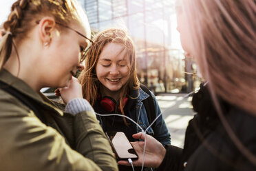 Teenage girl looking at friends listening music through mobile phone in city - MASF08848