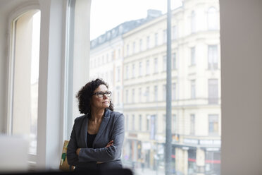 Thoughtful mature businessman looking through window while standing with arms crossed at office - MASF08868