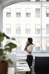 Seitenansicht einer reifen Frau, die mit ihrem Handy telefoniert und durch ein Fenster im Büro schaut - MASF08878