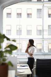 Side view of mature woman talking on mobile phone while looking through window at office - MASF08878