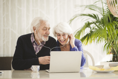 Fröhliches Seniorenpaar mit In-Ear-Kopfhörern bei Videotelefonie über Laptop im Pflegeheim, lizenzfreies Stockfoto