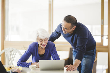 Cheerful mature man standing by mother looking at laptop on table in nursing home - MASF08921