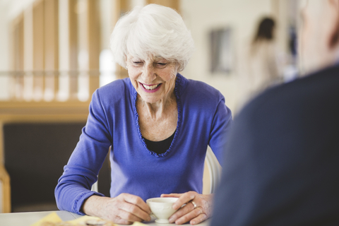 Ältere Frau lächelt bei einem Getränk am Tisch mit einem Mann in einem Pflegeheim, lizenzfreies Stockfoto