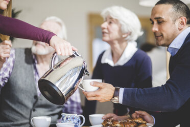 Abgeschnittene Hand einer Frau, die ein Getränk in eine von ihrem Vater gehaltene Tasse gießt, während die Großeltern am Tisch im Pflegeheim sitzen - MASF08940