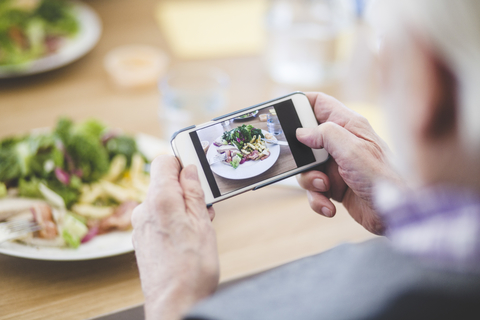Ausgeschnittenes Bild eines älteren Mannes, der mit seinem Mobiltelefon am Tisch Essen fotografiert, lizenzfreies Stockfoto