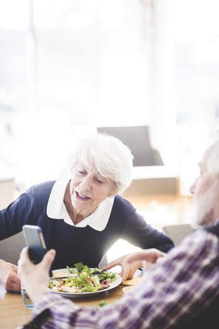 Älterer Mann zeigt Frau beim Mittagessen am Tisch im Pflegeheim sein Mobiltelefon, lizenzfreies Stockfoto