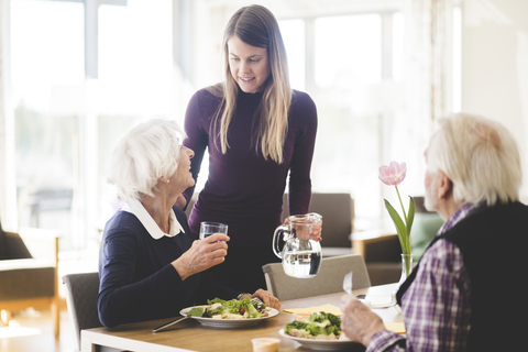 Junge Frau im Gespräch mit der Großmutter, während der Großvater am Tisch im Pflegeheim isst, lizenzfreies Stockfoto