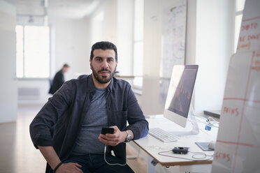 Portrait of male engineer holding mobile phone while sitting at computer desk in office - MASF08969