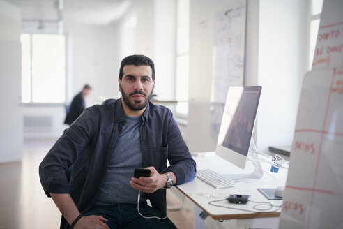 Porträt eines männlichen Ingenieurs, der ein Mobiltelefon hält, während er an einem Computertisch im Büro sitzt - MASF08969