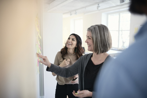 Mature female engineers smiling while reading adhesive notes stuck on glass in office stock photo