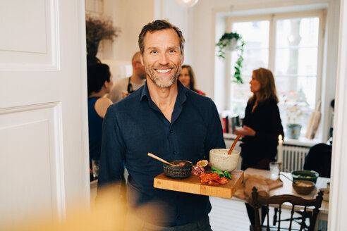 Porträt eines lächelnden reifen Mannes, der Essen in einem Serviertablett hält, während er auf einer Party vor Freunden steht - MASF09078