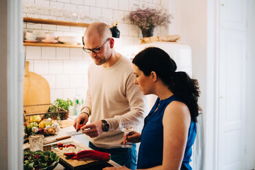 Woman with wineglass looking at male friend cutting pepper in kitchen - MASF09092