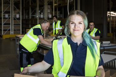 Porträt einer lächelnden Arbeiterin mit gefärbten Haaren, die in einem Vertriebslager vor Kollegen steht - MASF09126