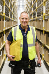 Portrait of smiling senior male worker holding package while standing on aisle at distribution warehouse - MASF09144