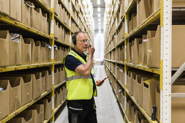 Senior male worker with digital tablet looking at packages on rack while talking through headset at distribution warehou - MASF09147