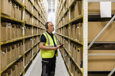 Side view of senior male worker with digital tablet looking at packages on rack while talking through headset at distrib - MASF09148